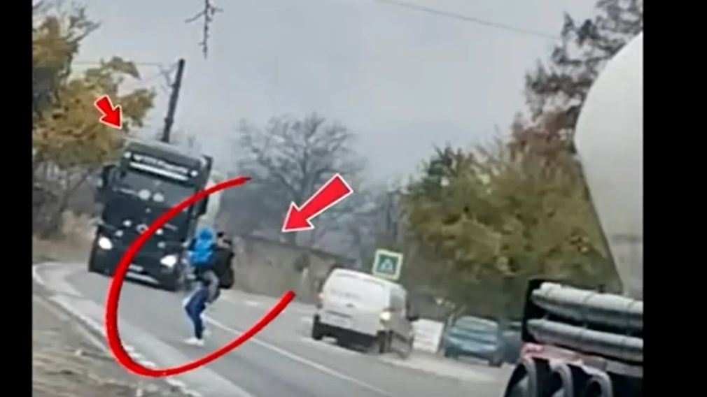 Imagine cu o mamă cu copilul în brațe, trecând printre două camioane